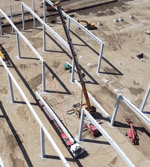 Transport agabaritic cu macara ridicând o grindă metalică pe șantier, operațiune de montaj structură industrială.