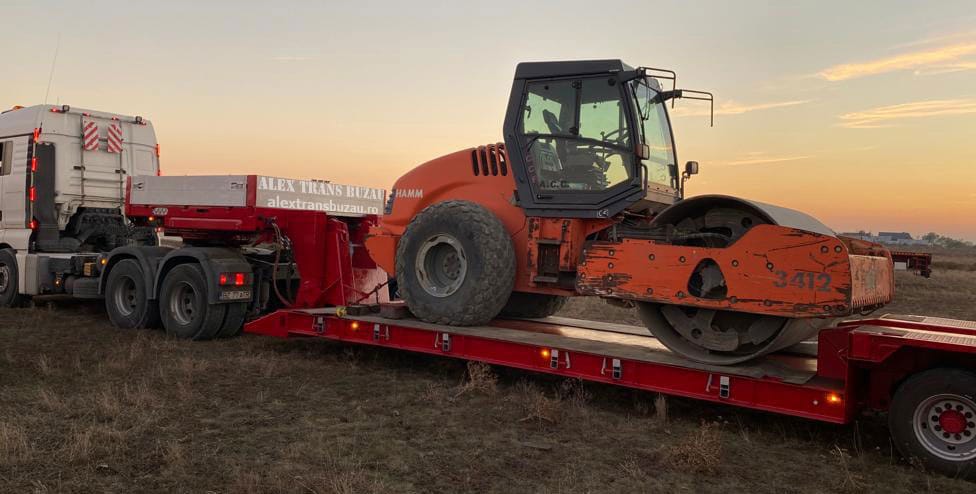 Camion cu semiremorcă low-loader roșie transport utilaje grele: compactator Hamm portocaliu pe teren neamenajat, monitorizare GPS activă