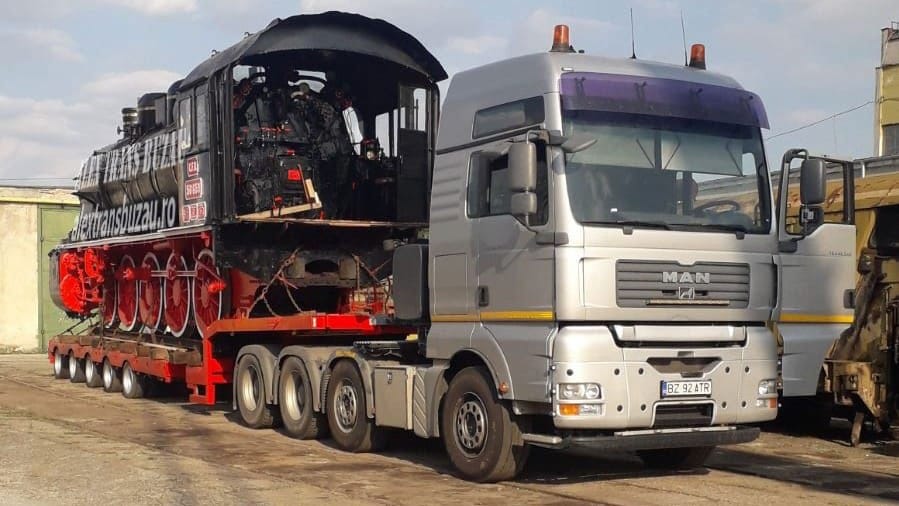Tractor MAN cu camion platformă transport utilaje grele, locomotivă istorică gri-roșie pe trailer low-loader Faymonville, stopuri reflectorizante
